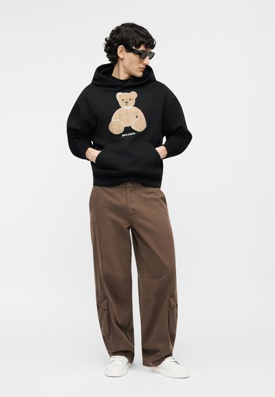 Persoon die een zwarte hoodie met teddybeerdessin draagt, bruine wijde cargobroek, witte sneakers en zwarte zonnebril, staand met de handen in de zakken.