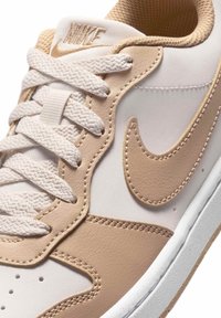 Κοντινή λήψη μπεζ και λευκού αθλητικού παπουτσιού Nike με λευκά κορδόνια, ραμμένο σύμβολο Nike και επενδεδυμένο γιακά.