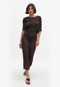 Brown draped top with short sleeves paired with a sheer brown skirt, featuring a smooth texture. Worn with dark sandals and a bangle.