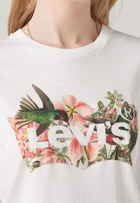 Weißes Baumwoll-T-Shirt mit einem bunten Blumenmotiv, das einen Kolibri zeigt, und dem "Levi's"-Text in fetten weißen Buchstaben in der Mitte.