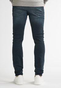 Persona vistiendo jeans ajustados azul oscuro y zapatillas blancas, vista desde atrás sobre un fondo blanco liso.