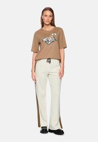 Braunes T-Shirt mit kurzen Ärmeln und einem bunten Grafikdesign eines Hundes, kombiniert mit hellbeigen Hosen mit Seitenschlitzen und einem Zugbandbund. Schwarze Schuhe.