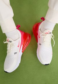 Nike Air Max 270 sneakers feature a cream upper, pink accents, and a large translucent red air sole. The design includes textured mesh and branding.