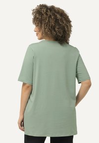 T-shirt verde chiaro a maniche corte con vestibilità rilassata, scollatura arrotondata e orlo sul retro più lungo. Texture liscia, design semplice senza decorazioni.