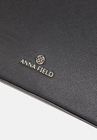 Musta teksturoitu pinta hienovaraisella kuvioinnilla, jossa on kullan kohokuvioitu "ANNA FIELD" -logo ja kukkakuvio brändäystä varten.