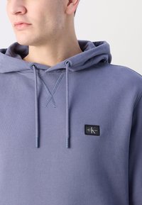 Felpa con cappuccio viola realizzata in un tessuto morbido, dotata di un piccolo patch con logo nero, cordini regolabili e dettagli a coste sul colletto.