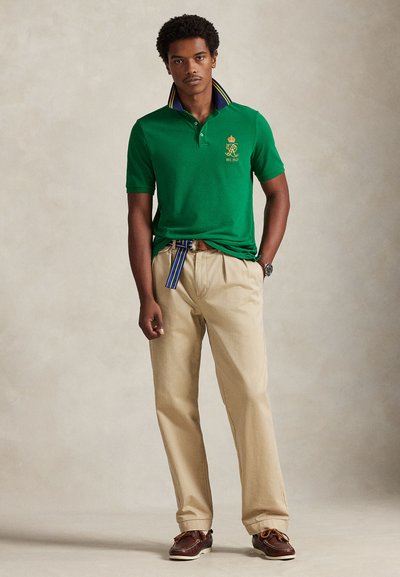 Πράσινο πόλο με μπλε και κίτρινο ριγέ γιακά, μπεζ παντελόνι και καφέ παπούτσια. Σημαντικό λογότυπο κορόνας και λεπτομέρεια ημερομηνίας ίδρυσης στο πουκάμισο.