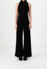 Mono de terciopelo negro con un alto cuello y una cremallera frontal. Presenta piernas anchas y un cuerpo ajustado, textura suave y sin patrones visibles.