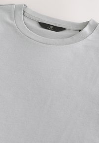 T-shirt gris clair à col rond avec une étiquette noire portant la mention « NEXT » à l'intérieur du col.