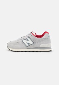 Šedá teniska New Balance s bílým logem "N", červeným vnitřním podkladem a černou podrážkou, zobrazená v profilu na bílém pozadí.