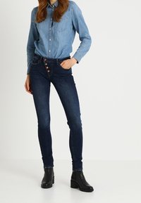 Mörkblå slim fit jeans med knapprad och femfickdesign, i kombination med en ljus denimskjorta som har krage och två bröstfickor.