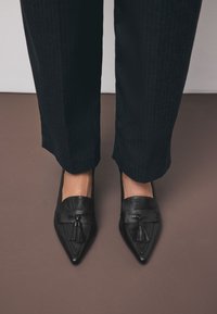 Mocassins en cuir noir à bouts pointus, ornés de pampilles décoratives et de détails de couture. Portés avec un pantalon à rayures fines foncé.