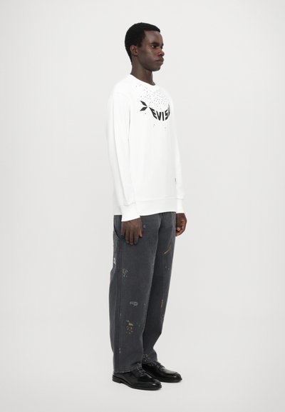 Hvid langærmet skjorte med sort logo, parret med mørkeblå bukser med farverige mønstre og stikninger. Sorte sko fuldender outfittet.