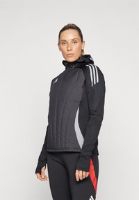 Veste athlétique noire avec un devant matelassé, des accents latéraux gris et trois bandes blanches sur les épaules. Inclut une capuche et des trous pour les pouces.