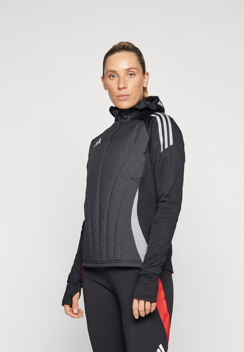 Veste athlétique noire avec un devant matelassé, des accents latéraux gris et trois bandes blanches sur les épaules. Inclut une capuche et des trous pour les pouces.