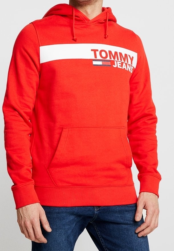 Sweat à capuche rouge avec une bande blanche, texte "Tommy Jeans" et logo sur la poitrine, porté avec un jean bleu par une personne debout.