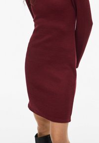 Abito bordeaux a coste con maniche lunghe, silhouette aderente e orlo al ginocchio. Tessuto liscio. Nessun motivo o dettaglio visibile.