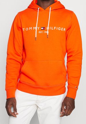 Mann trägt eine leuchtend orangefarbene Tommy Hilfiger Kapuzenjacke mit weißen Kordeln und weiße Hose, steht vor einem schlichten Hintergrund.
