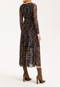 Eine Frau steht mit dem Rücken zur Kamera, trägt ein langes, gemustertes Kleid in Erdtönen und beige Ankle Boots mit hohen Absätzen.