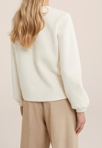 Nainen, jolla on pitkät vaaleat hiukset, käyttää teksturoitua valkoista pitkähihaista puseroa ja beigejä leveälahkeisia housuja, kuvattuna takaapäin neutraalia taustaa vasten.