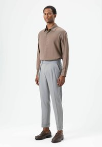 Chemise marron à manches longues avec col pointu, pantalon plissé gris, et mocassins foncés. Présente un tissu lisse et une coupe ajustée.