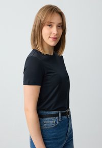T-shirt nero a maniche corte con scollatura rotonda, indossato con jeans blu a vita alta e una cintura nera. Tessuto morbido, vestibilità casual.
