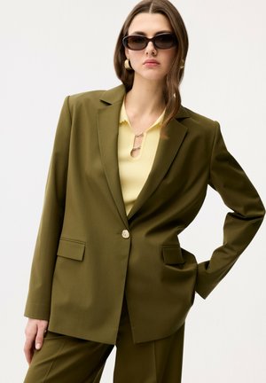 Frau trägt übergroßen olivgrünen Blazer und Hose mit gelbem Oberteil, dunkler Sonnenbrille und goldenen Creolen, posiert mit der Hand in der Hüfte.