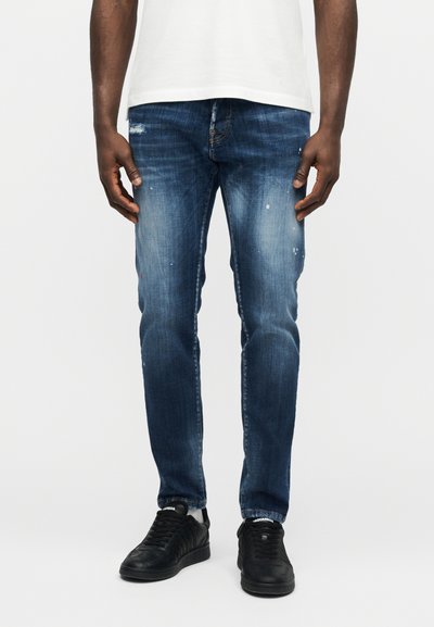 Homme portant un jean slim bleu délavé avec des effets d'usure subtils et des baskets noires, debout devant un fond blanc uni.
