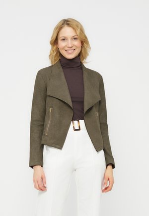 Mosolygó szőke nő olívazöld dzsekit visel sötét garbó fölött, fehér magasderekú nadrágot barna övcsattal.