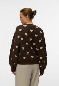 Hnědý pletený svetr s světle béžovými srdíčkovými vzory, dlouhými rukávy, volným střihem a lemováním na rukávech a spodním lemu, nošený přes světlé kalhoty.