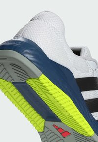 Zapatilla deportiva gris y blanca con parte superior texturizada, acentos en azul marino, franjas negras y suela vibrante amarilla con un patrón de taco.