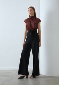 Bruine satijnen top met een hoge ruches kraag, gecombineerd met zwarte wijde broek met een strikceintuur. Model draagt zwarte hoge hakken sandalen.