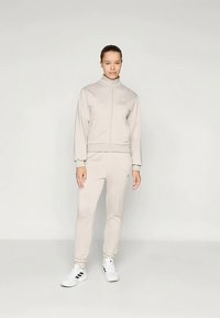 Ανοιχτό μπεζ αθλητική φόρμα με ζακέτα με φερμουάρ και ταιριαστά στενά παντελόνια, κατασκευασμένη από λείο ύφασμα. Λευκά αθλητικά παπούτσια με μαύρες ρίγες αντίθεσης.