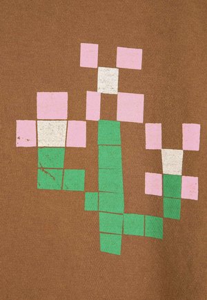 Mujer con una camiseta marrón de gran tamaño con estampados pixelados de flores, una falda beige envolvente de talle alto y zapatos crema sin cordones.