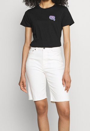 Mujer con una camiseta negra ajustada con un pequeño gráfico púrpura en el pecho y pantalones cortos de mezclilla blancos hasta la rodilla con dobladillos deshilachados.