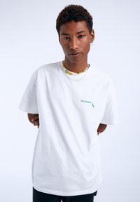 Bílá bavlněná tričko s krátkým rukávem, na levé straně hrudi s nápisem "DREAM GOOD TIMES" v zelené barvě, uvolněný střih, jednoduchý design.