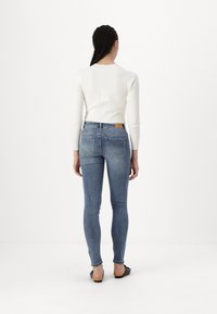 Jeans de denim azul ajustados con una cintura plana, dos bolsillos traseros y una parche de cuero amarillo. Combinados con una camiseta de manga larga blanca de canalé.