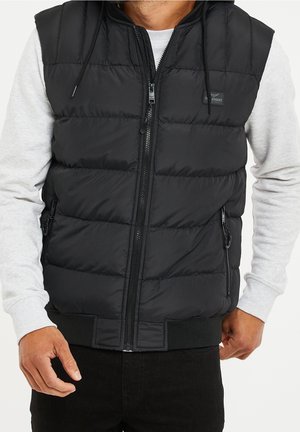 Mann trägt schwarze, gesteppte ärmellose Weste mit Reißverschluss über hellgrauem Langarmshirt und schwarzen Hosen, Hände seitlich sichtbar.