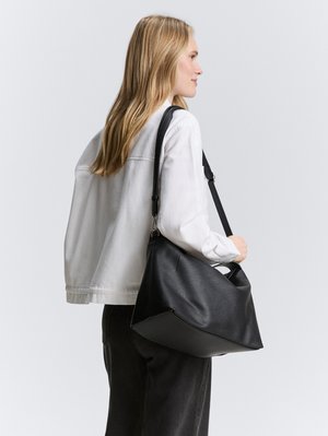 Femme aux longs cheveux blonds portant une veste blanche et un pantalon noir, portant un grand sac à bandoulière en cuir noir sur son épaule droite.