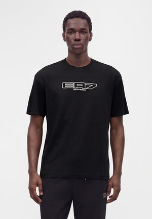 Άνδρας που φοράει μαύρο T-shirt EA7 Emporio Armani και μαύρο παντελόνι μπροστά σε λευκό φόντο, κοιτάζοντας μπροστά με ουδέτερη έκφραση.