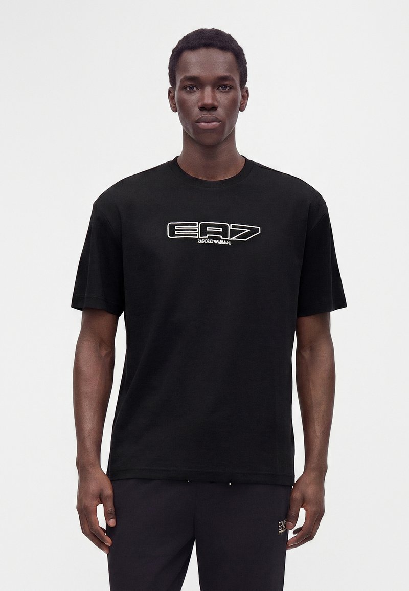 Άνδρας που φοράει μαύρο T-shirt EA7 Emporio Armani και μαύρο παντελόνι μπροστά σε λευκό φόντο, κοιτάζοντας μπροστά με ουδέτερη έκφραση.