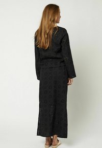 Zwart tweedelig outfit met lange mouwen en een maxirok, voorzien van bloemenoogjespatronen en een zijsplit tot halverwege het bovenbeen.