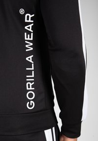 Zwarte jas met witte verticale strepen en "GORILLA WEAR" in witte letters gedrukt op de linkermouw. Gemaakt van gladde stof, met elastische manchetten.