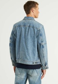 Chaqueta de mezclilla azul claro con un diseño clásico, cierre de botones y detalles de costura en el pecho. Presenta una tela ligeramente texturizada y un logo en la parte posterior.