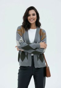 Kobieta uśmiechająca się ze złożonymi ramionami, ubrana w szary wzorzysty kardigan, biały top, czarne spodnie i nosząca brązową torebkę na ramię.