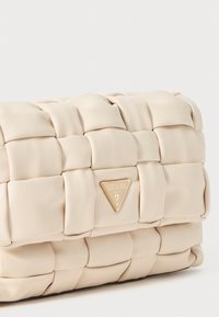 Beige műbőr táska steppelt mintázattal, elöl egy háromszögletű arany logó táblával. Kompakt téglalap alakú.