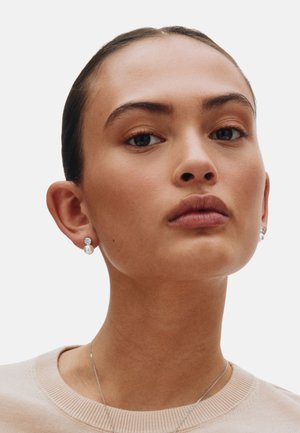 Orecchini d'argento con gemme tonde e pendente di perla, indossati da una persona con espressione neutra. È visibile un maglione beige chiaro.