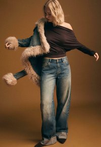 Giacca di jeans con colletto in pelliccia, abbinata a un top marrone senza spalline e jeans larghi blu, su uno sfondo marrone.