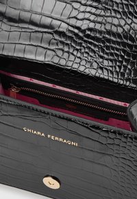 Svart väska i krokodilmönster med guldklämma och Chiara Ferragni-logotyp, som visar ett rosa tyginteriör och innerfickan med dragkedja.