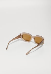 Heksagonale solbriller med en transparent lys rosa ramme og brune tonede linser. Slanke, buede stenger med metalldetaljer.